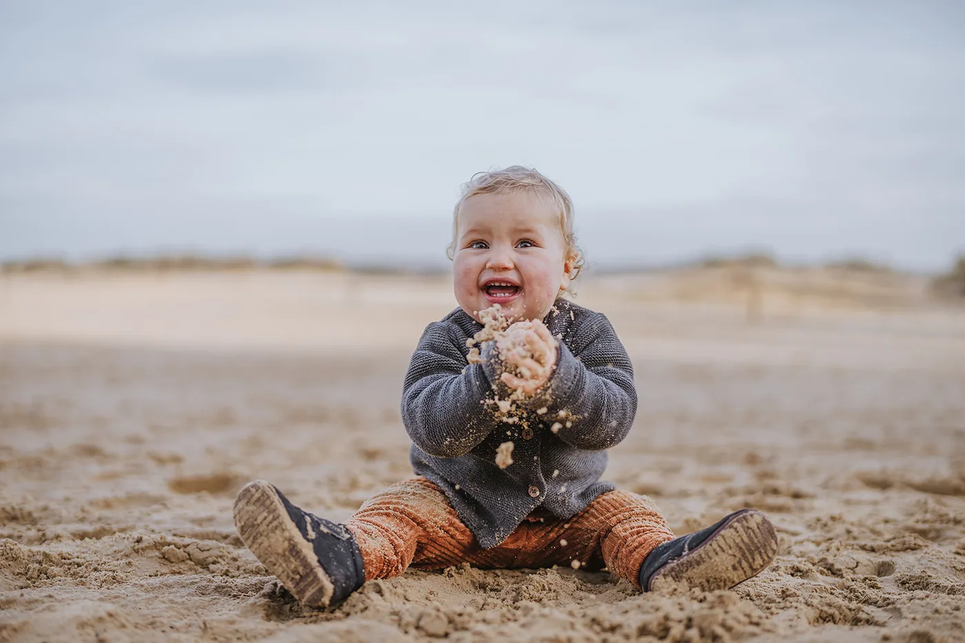 Peuter zit midden in het zand te lachen, handen vol zand