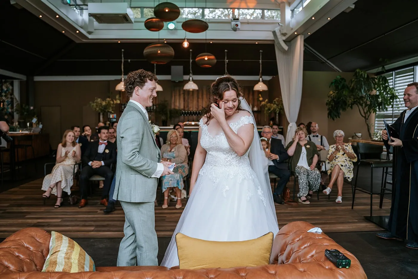 Bruid veegt een traan weg tijdens de geloften, de bruidegom kijkt lachend toe en achter hen zitten de gasten in de ceremonieruimte