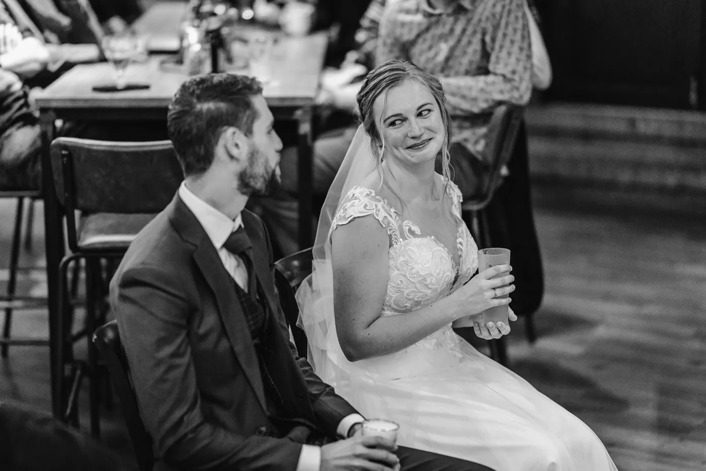 Bruidspaar zit naast elkaar te lachen tijdens de speeches, in zwart-wit