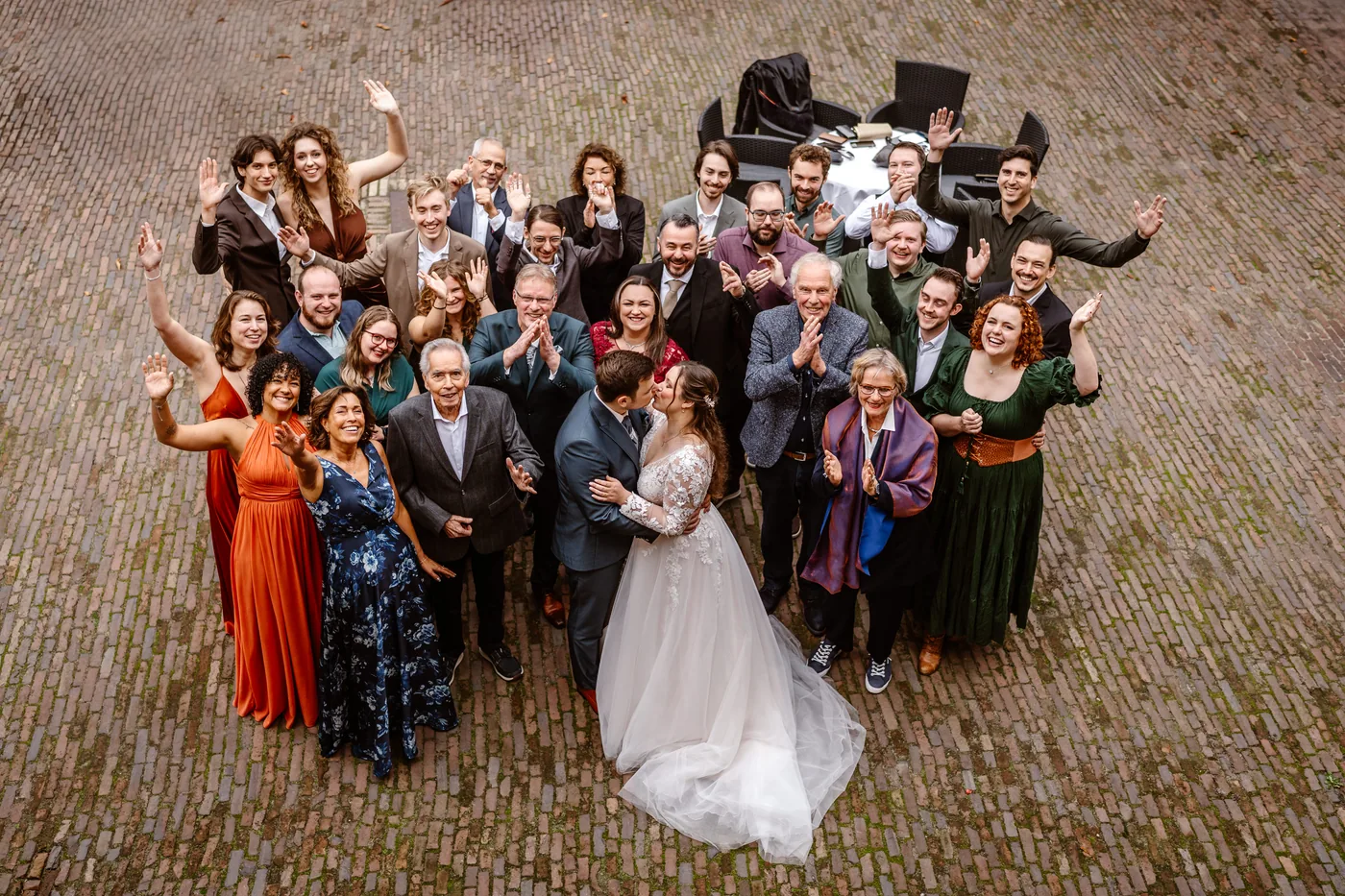 Groepsfoto van bovenaf op een geplaveide binnenplaats: het bruidspaar kust elkaar midden in de kring, de gasten eromheen zwaaien en klappen omhoog naar de camera
