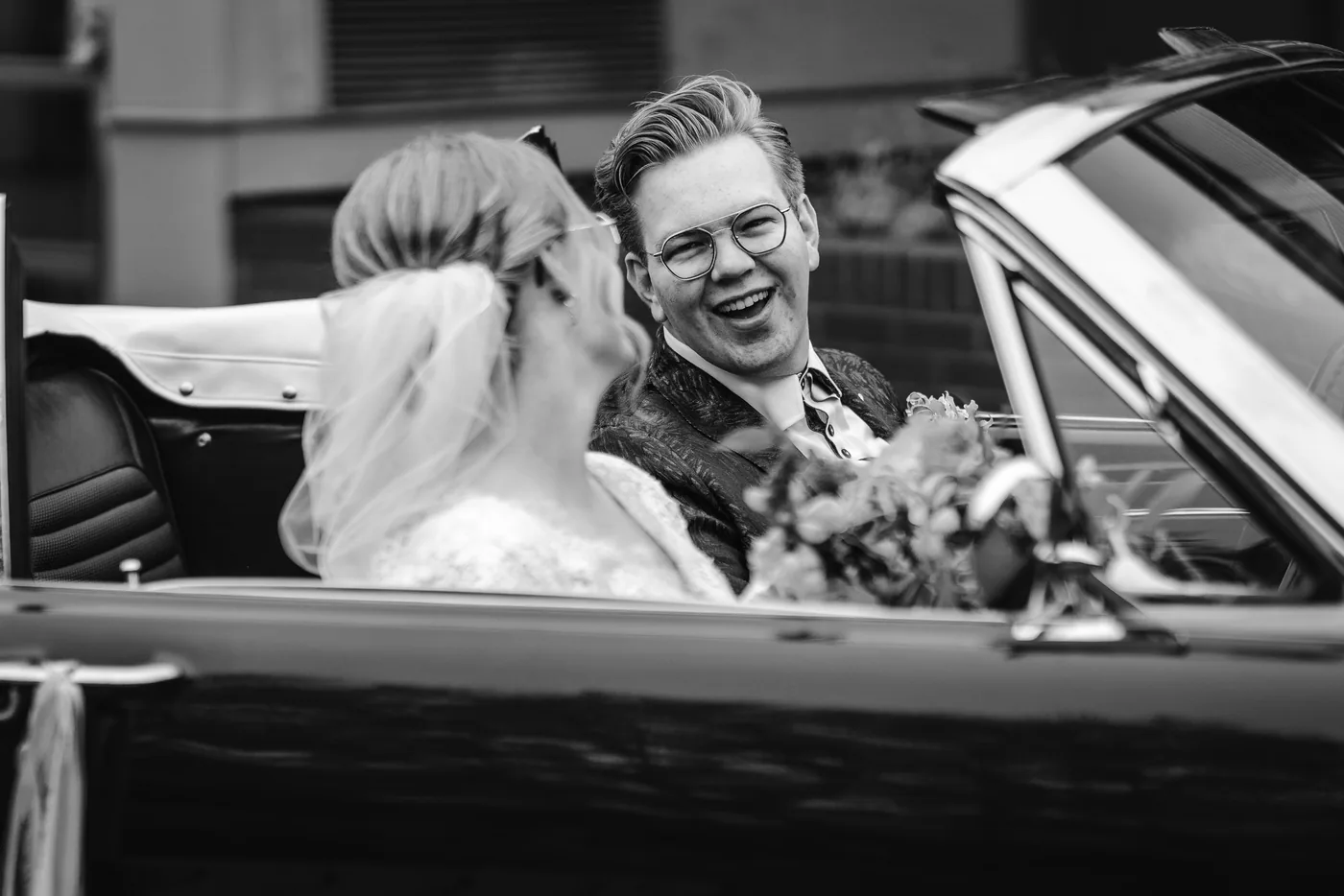 Bruid en bruidegom lachen samen in een open auto onderweg naar de trouwlocatie